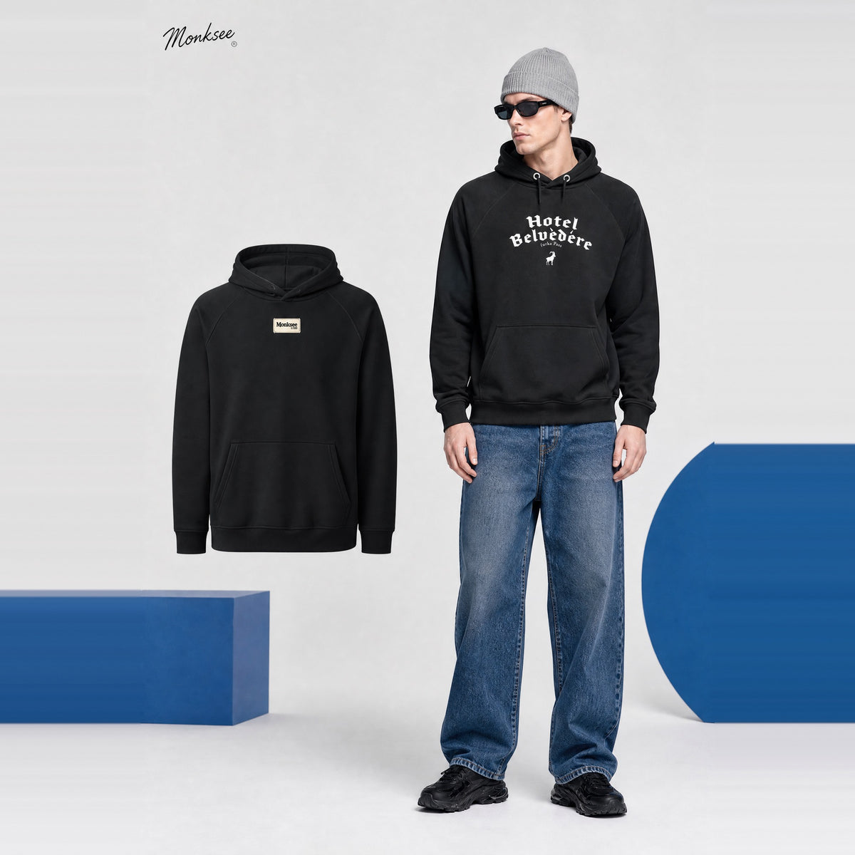 Model wearing the black Monksee Hotel Belvedere Furka Pass eco raglan hoodie with jeans and a grey beanie, shown next to the hoodie laid flat