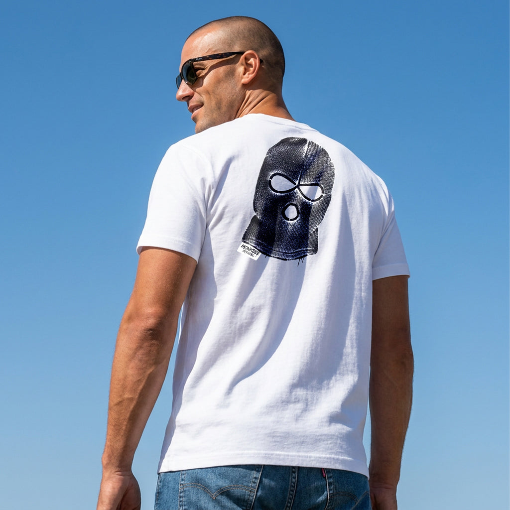 Man wearing a white t-shirt with a balaclava with a Monksee label on the back against a clear blue sky.