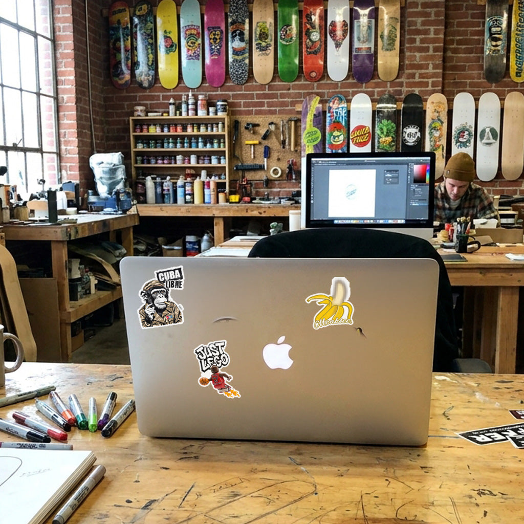 A silver Apple MacBook laptop sits open on a wooden worktable in a busy skate shop. The laptop is decorated with several stickers, including a 'Cuba Libre' sticker, a banana sticker, and a 'Just Lego' sticker.