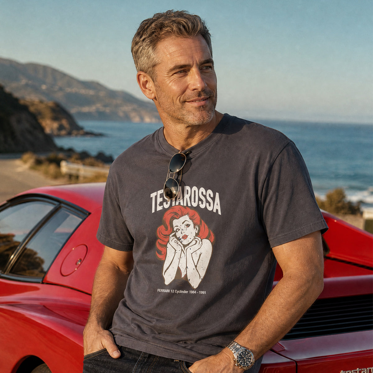 Man wearing the Monksee Testarossa garment-dyed t-shirt in navy, leaning on a red Ferrari Testarossa on a coastal road at golden hour