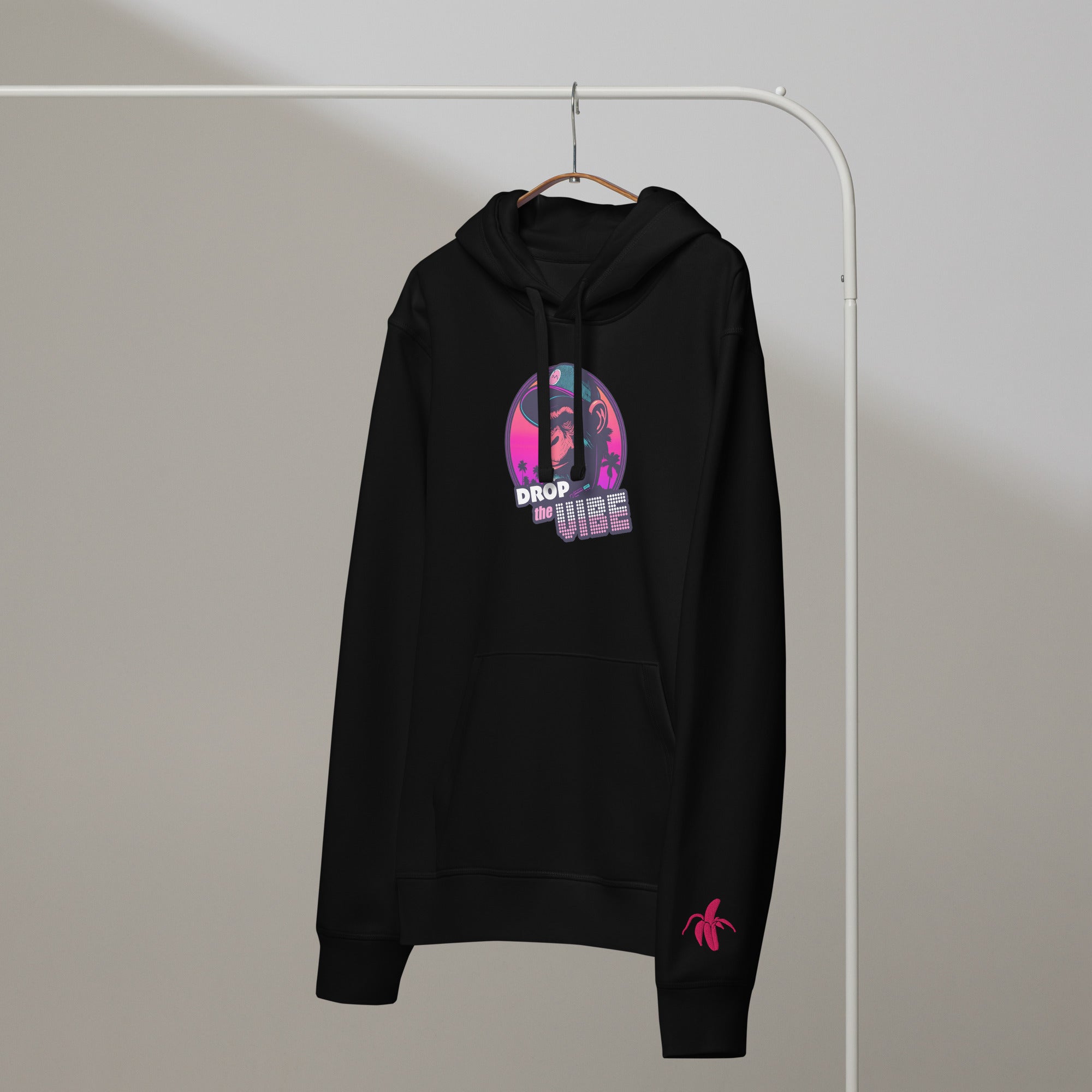 A black Monksee "Drop the Vibe" hoodie hanging on a rack against a white background. It features a neon graphic of a cap-wearing monkey against a sunset with palm trees, with "DROP the VIBE" in bold pink and white text. A small pink Monksee banana logo is on the left sleeve