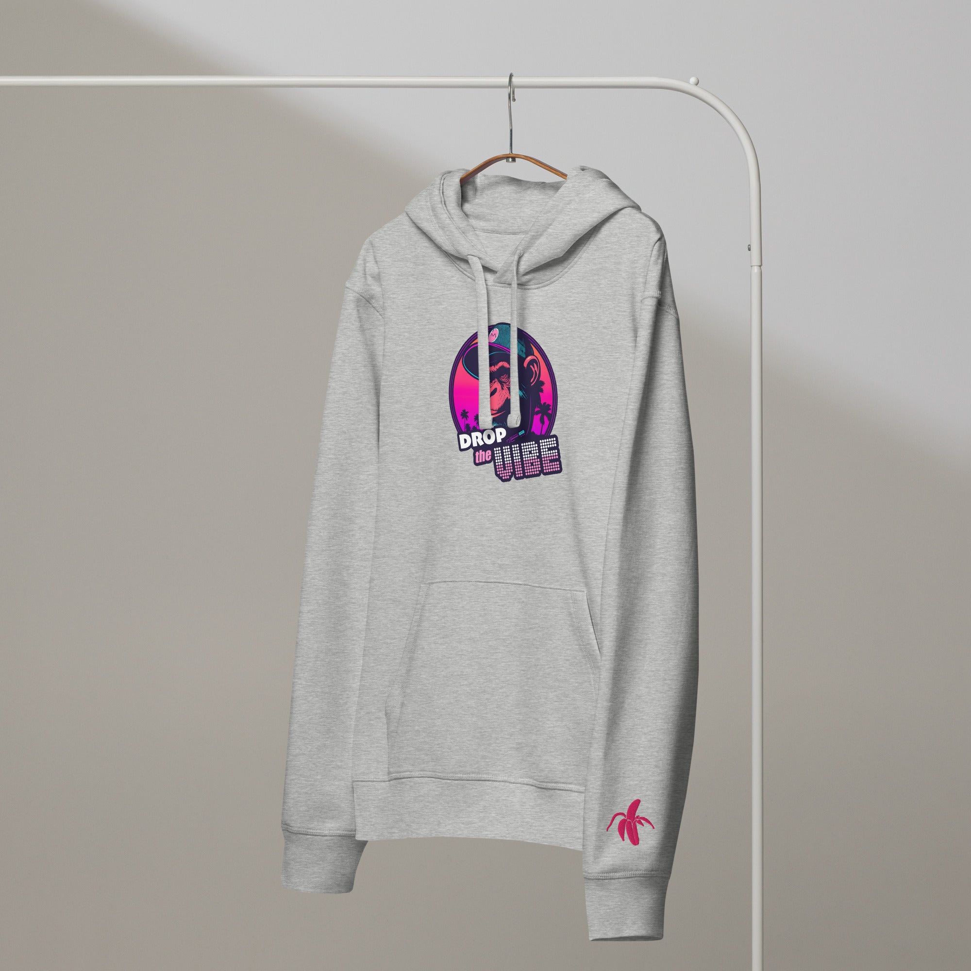 A grey Monksee "Drop the Vibe" hoodie hanging on a rack against a white background. It features a neon graphic of a cap-wearing monkey against a sunset with palm trees, with "DROP the VIBE" in bold pink and white text. A small pink Monksee banana logo is on the left sleeve