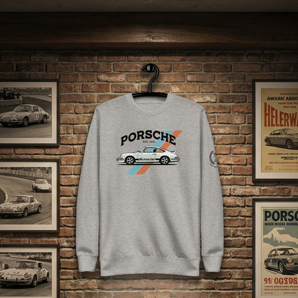 A light gray crewneck sweatshirt hanging on a black hanger against a brick wall, featuring a graphic print of a white Porsche 911 with orange and blue stripes and the text 'PORSCHE EST 1931'. Framed vintage car posters are visible on the brick wall background.