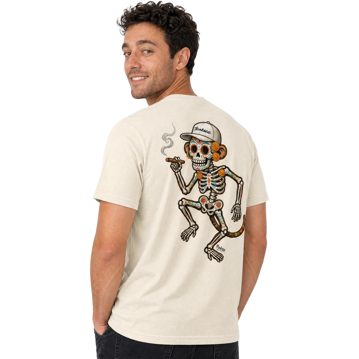 Model looking over his shoulder in the Monksee El Mono Muerto garment-dyed heavyweight t-shirt in Ivory, back view, showing the Día de los Muertos sugar-skull monkey skeleton print with marigolds, painted bones, a white Monksee cap and a smoking cigar