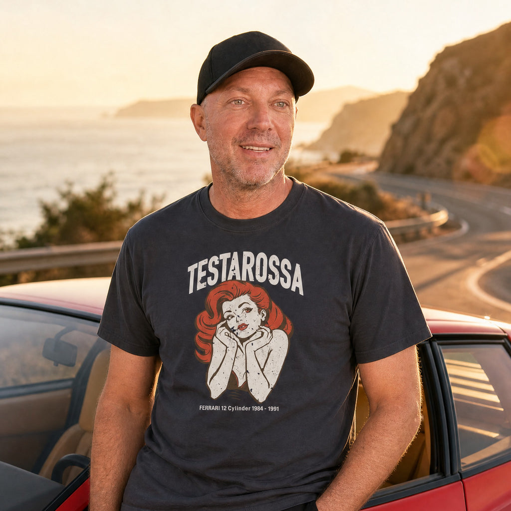 Man in a navy Monksee Testarossa garment-dyed t-shirt leaning on a red Ferrari Testarossa on a coastal road at sunset