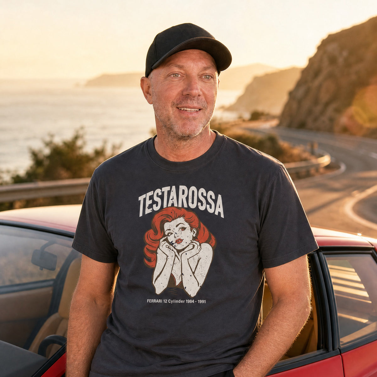 Man in a navy Monksee Testarossa garment-dyed t-shirt leaning on a red Ferrari Testarossa on a coastal road at sunset